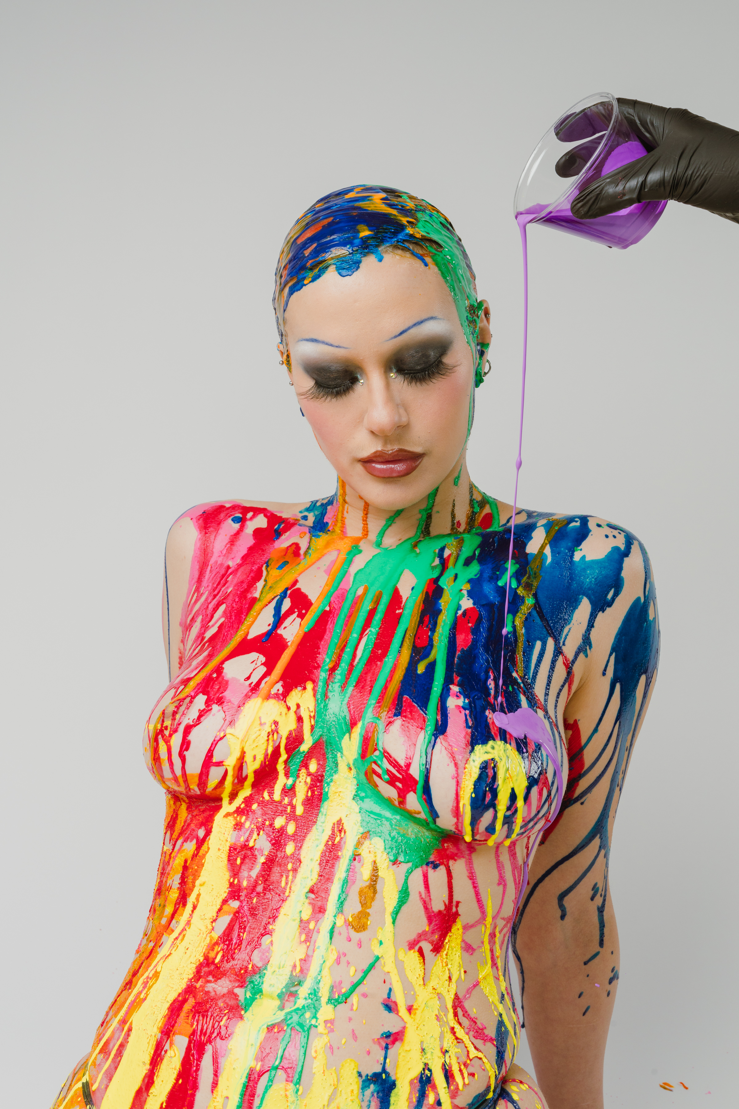 A disembodied hand in a black nitrile glove drips purple paint on the subject of the photoshoot. Their darkened lips, long lashes, intense grey eyeshadow that blends into white as it crosses into the brow area, and thin blue eyebrows that almost appear to be on a cloud formed by the white eyeshadow are featured prominently.